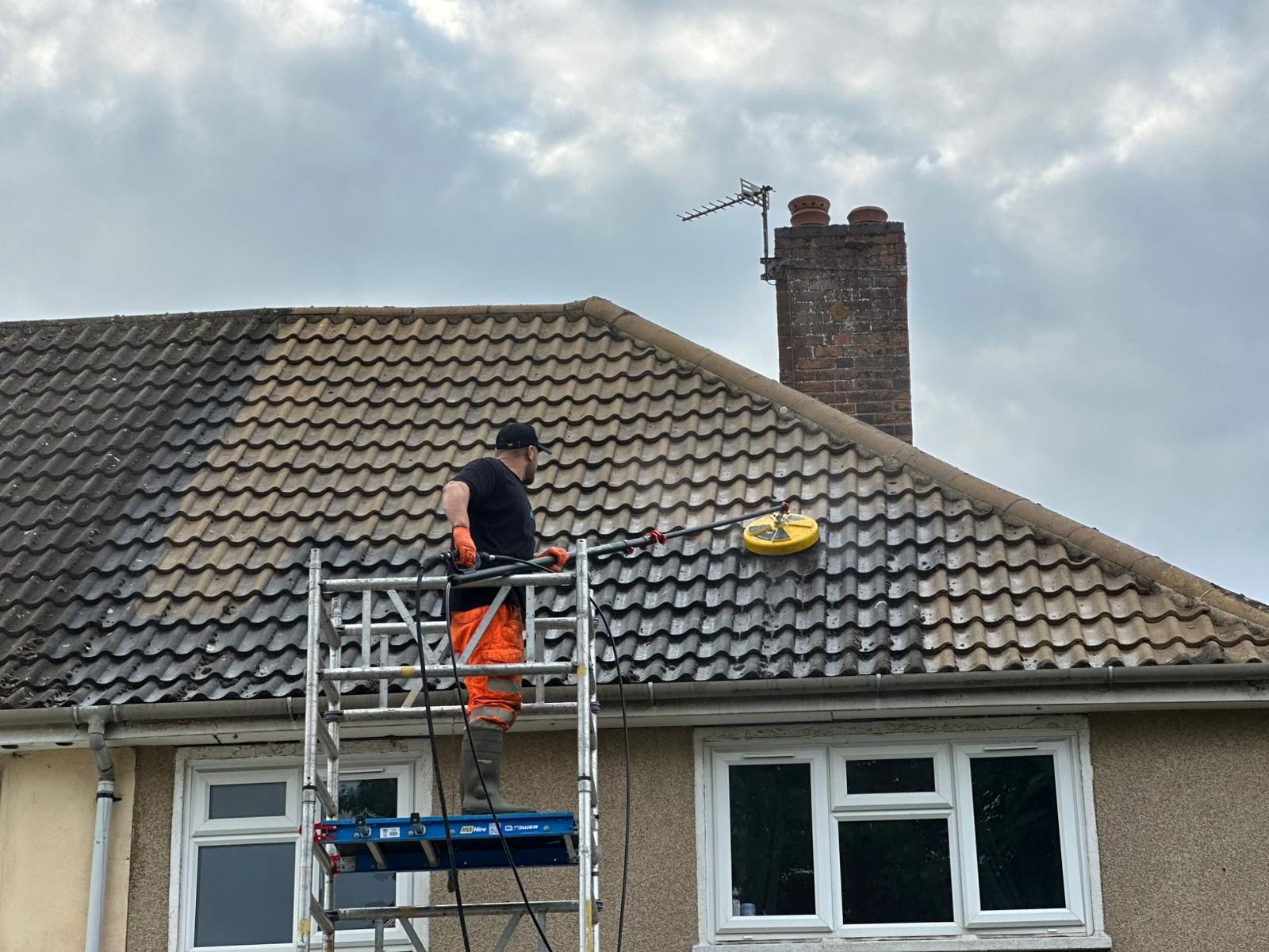 Technician on scaffolding using a yellow circular roof cleaning tool on a residential roof, showing a clear difference between cleaned and uncleaned tiles